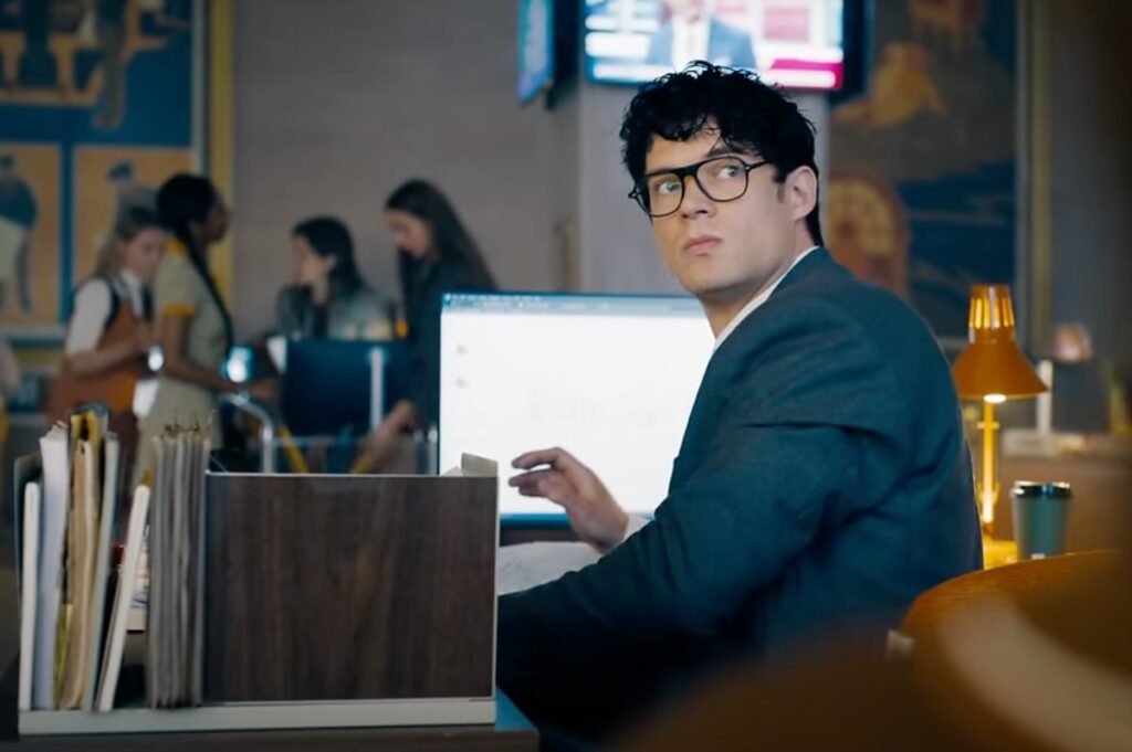 Young man with curly dark hair and glasses sitting at a desk in a newsroom.