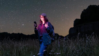 Photo illustration shows a woman holding a phone in a remote area, with dots in the sky to represent satellite service.