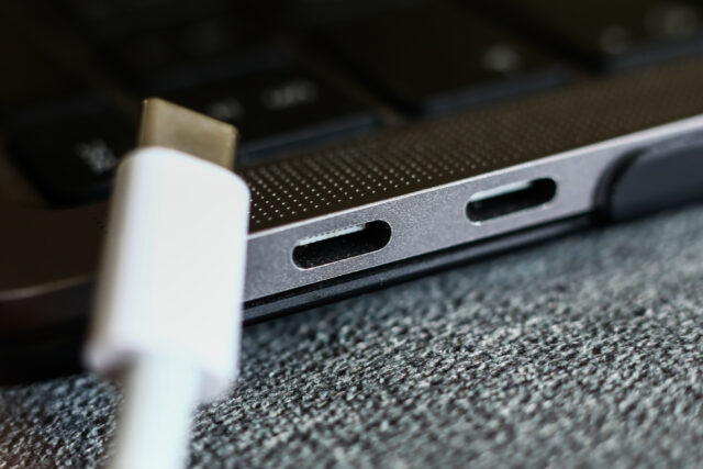 A USB-C plug in front of a laptop or other device with two USB-C ports showing, against a gray cloth background.