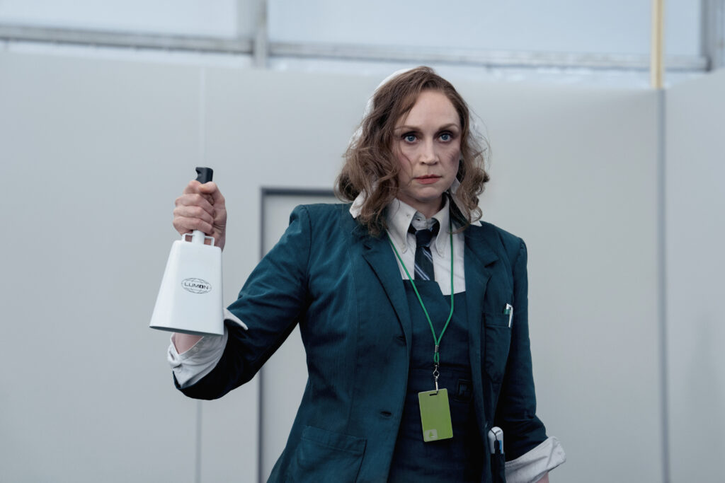 A disheveled woman carries a cowbell in a corporate office