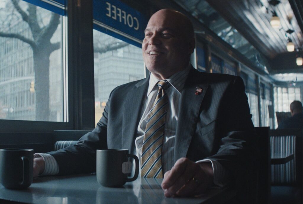 VInvent D'Onofrio as Wilson Fisk, also smiling in a suit sitting in a diner