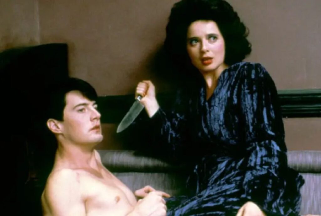 half naked man lying down with a woman in a blue robe holding a knife on top of him