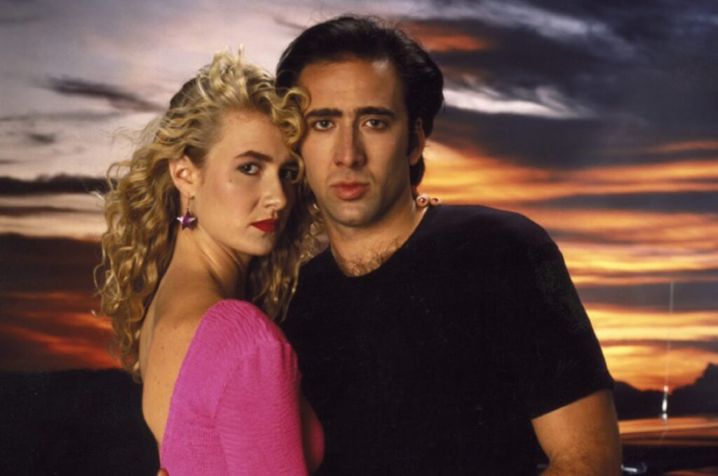 blonde woman in hot pink dress standing close to brooding dark haired man in a black t-shirt