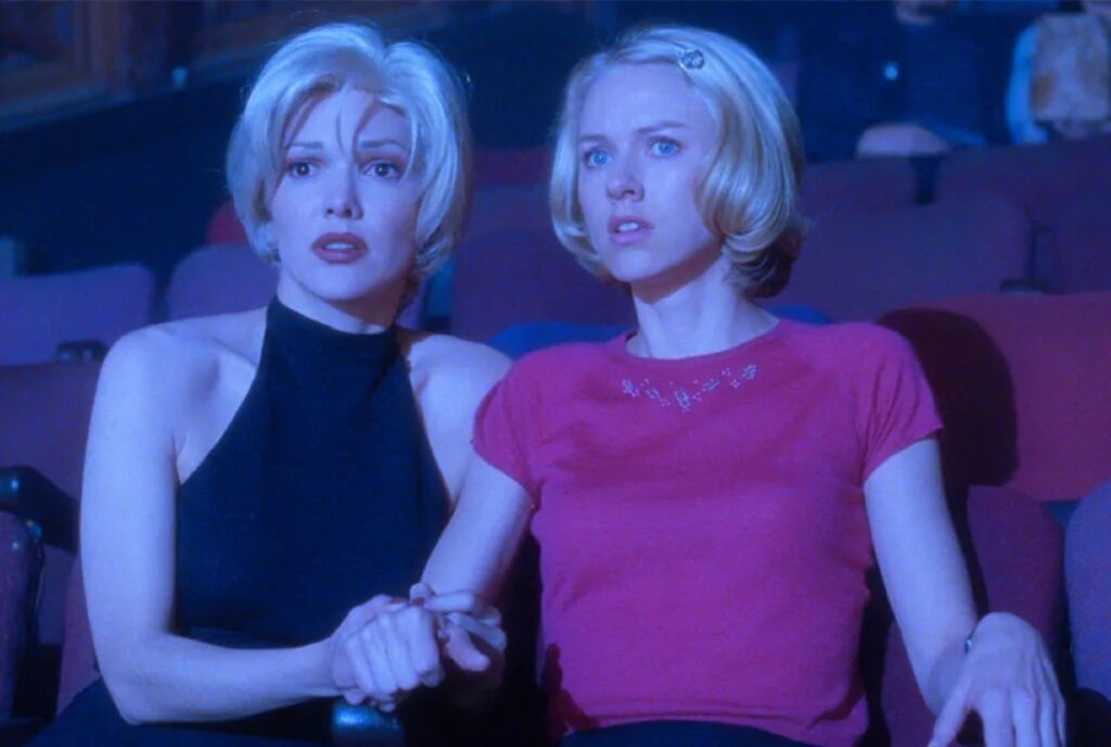 two similar-looking women with short blonde hair, one in a black top, one in a hot pink top, holding hand sin a movie theater