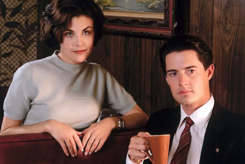 dark haired woman in light gray sweater sitting next to man in suit holding a coffee cup.