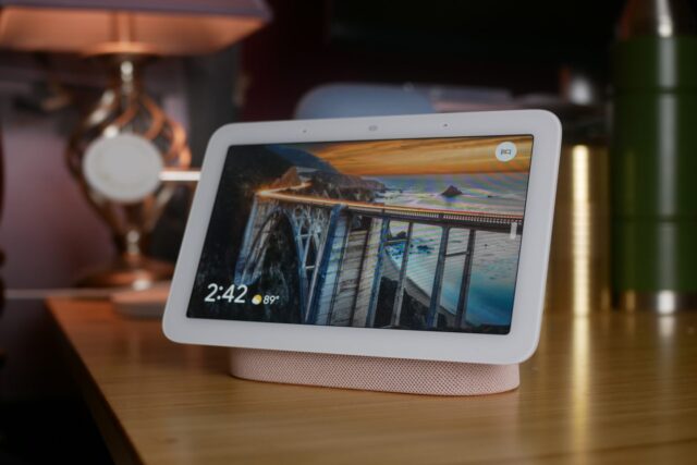 Nest Hub second generation, on a nightstand with a bamboo top and dim lamp in the near background.