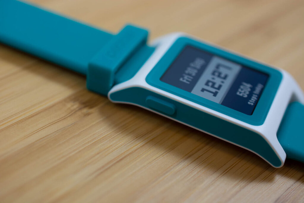 Pebble watch showing a text watchface (reading 12:27 p.m.), with greenh silicone band and prominent side button.