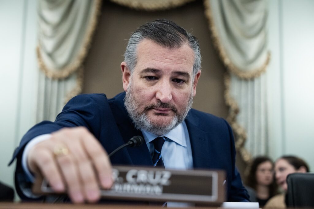 Senator Ted Cruz at a Senate committee hearing, sitting in his seat and using his hand to move a nameplate that says 