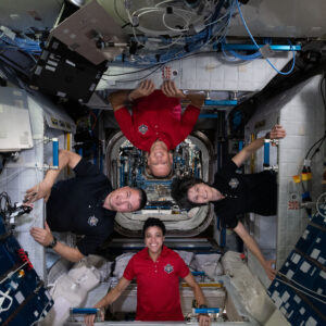 Image of four people, each standing, but on different sides of the ISS such that all of their heads are in the center of the photo. There are racks of equipment on most of the walls nearby.