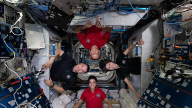 Image of four people, each standing, but on different sides of the ISS such that all of their heads are in the center of the photo. There are racks of equipment on most of the walls nearby.