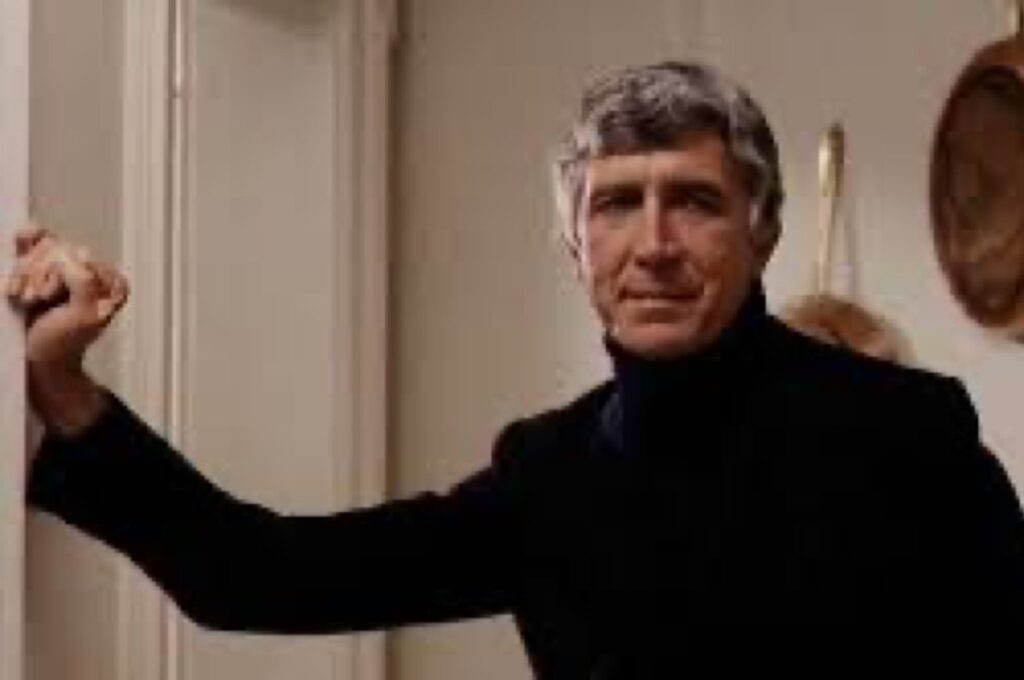 silver-haired man in black turtleneck with one hand on the door jamb