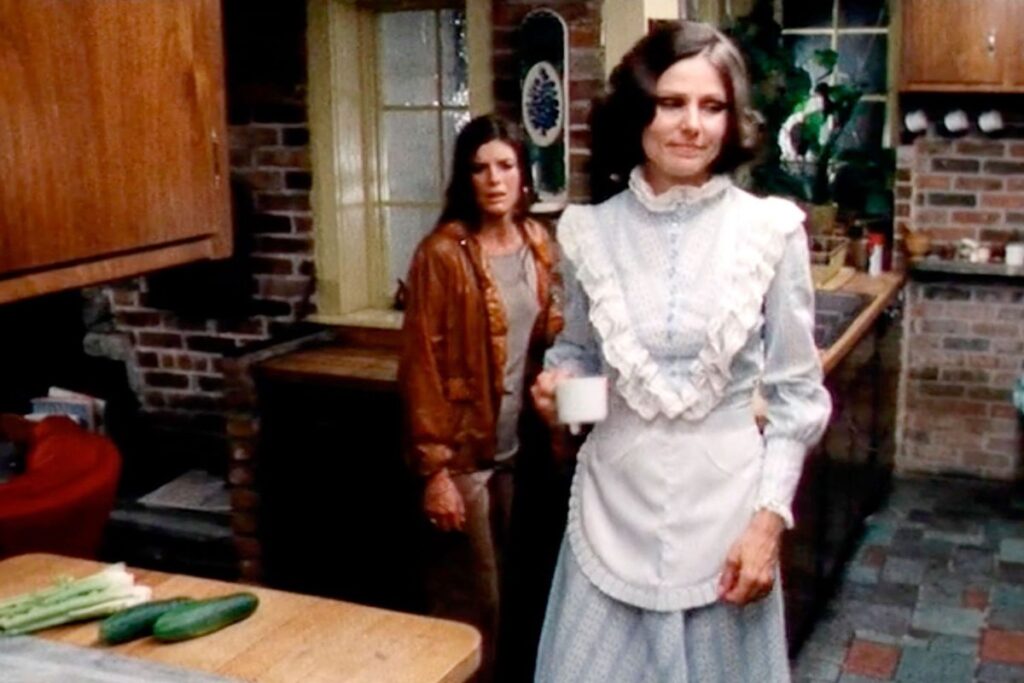 Brunette woman in apron and prairie dress in the kitchen holding a coffee cup while another woman looks on from the background