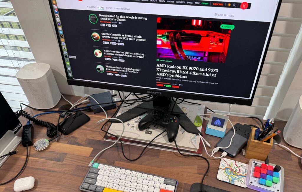 Wooden desk with a monitor opened to Ars Technica homepage, a keyboard, mouse, small ESP32 Home Asssistant voice device, Anker multi-charger, Figma keypad device, a MacBook, and too many cables.