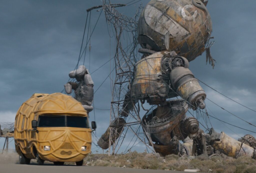 yellow peanut shaped van driving past giant ruined robot.