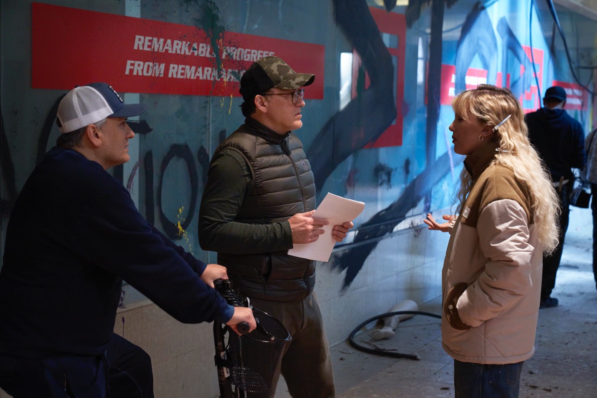 two men in baseball caps directing a young blonde woman in a yellow parka on a movie set