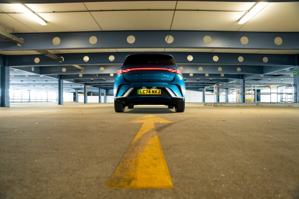 A BYD Dolphin seen from behind in a parking structure. A large yellow arrow painted on the floor points at the car