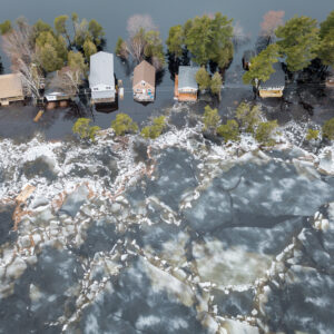 Ice forms near a row of trees near a set of houses with flooded yards, as seen from the air.