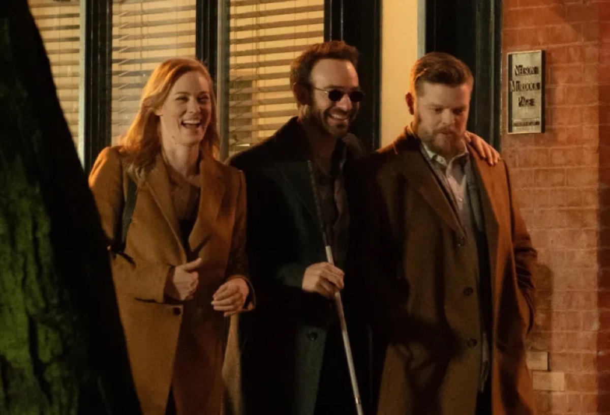 blonde woman, blind man, and pudgy bearded man talking arm in arm down the street smiling