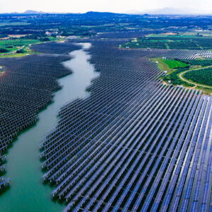 A large solar installation, with panels encroaching on a body of water in the center from both banks. Many panels are installed on stanchions in the water.