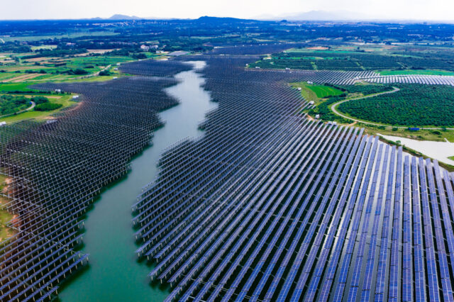 A large solar installation, with panels encroaching on a body of water in the center from both banks. Many panels are installed on stanchions in the water.