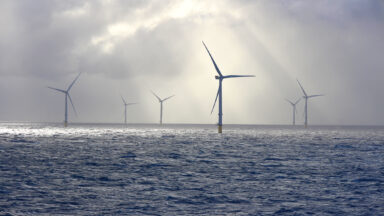 A series of tall wind turbines in the ocean, illuminated by beams of sunlight filtering through baps of heavy clouds.