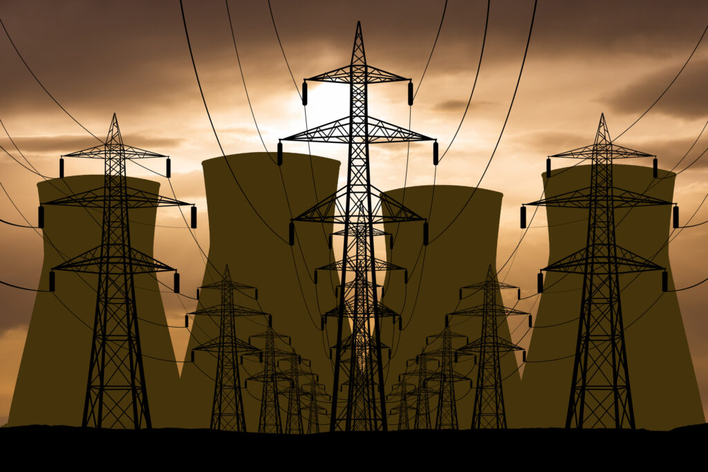 An orange, cloudy sky backlights a set of electrical wires on large pylons, leading away from the cooling towers of a nuclear power plant.