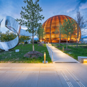 Image of a grassy area with an abstract metal sculpture on the left, and a wooden sphere on the right.