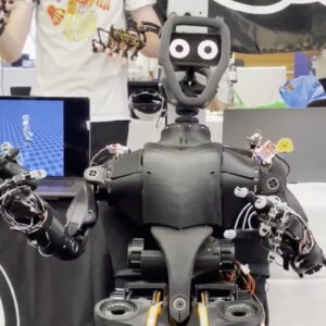 A humanoid robot sits in a chair, with a telepresence operator behind it