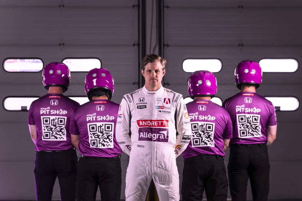 A racing driver stands in front of four mechanics, who are facing away from him. The mechanics have QR codes on the back of their shirts.