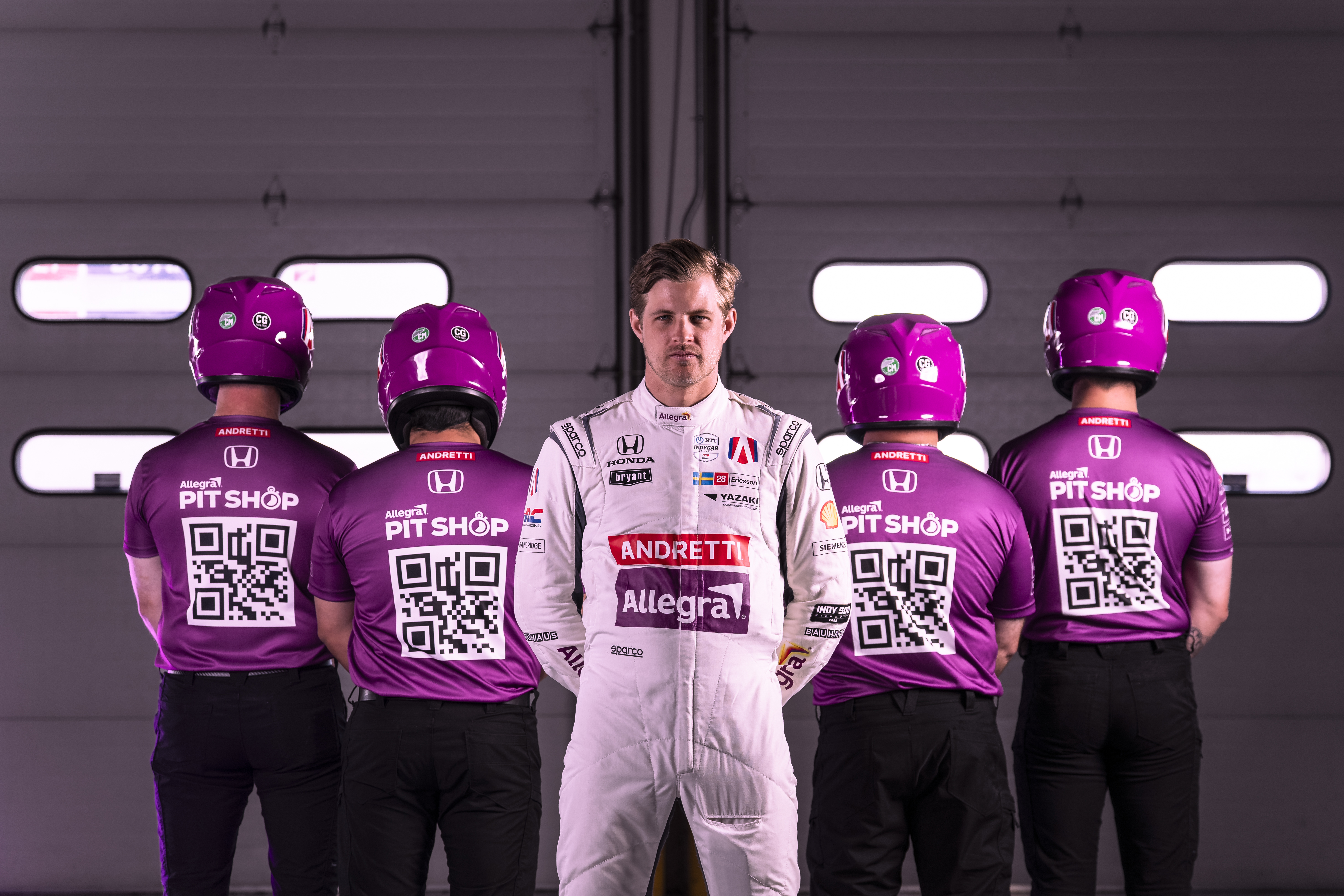 A racing driver stands in front of four mechanics, who are facing away from him. The mechanics have QR codes on the back of their shirts.