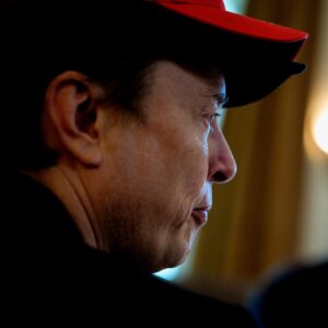 Elon Musk photographed from the side while sitting in a meeting and wearing a red Trump hat.
