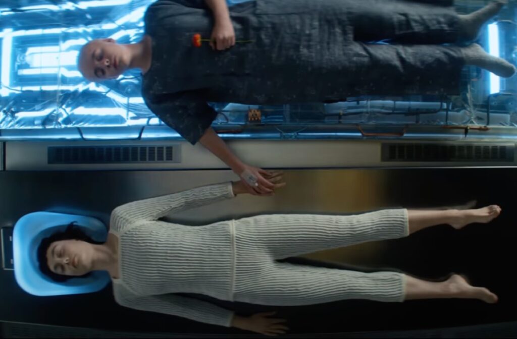 young girl with shaved head lying on medical bed next to another young woman dressed all in white. The two are holding hands.