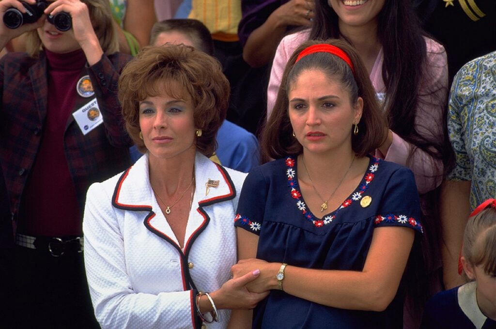 Two women in early 70s garb holding hands among a crowd in the grandstands