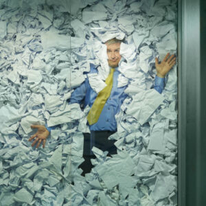 A businessman is trapped in his glass office by a surplus of discarded ideas on paper . His colleague in the next office is working more efficiently and is oblivious to him being trapped , as is a passing female office worker