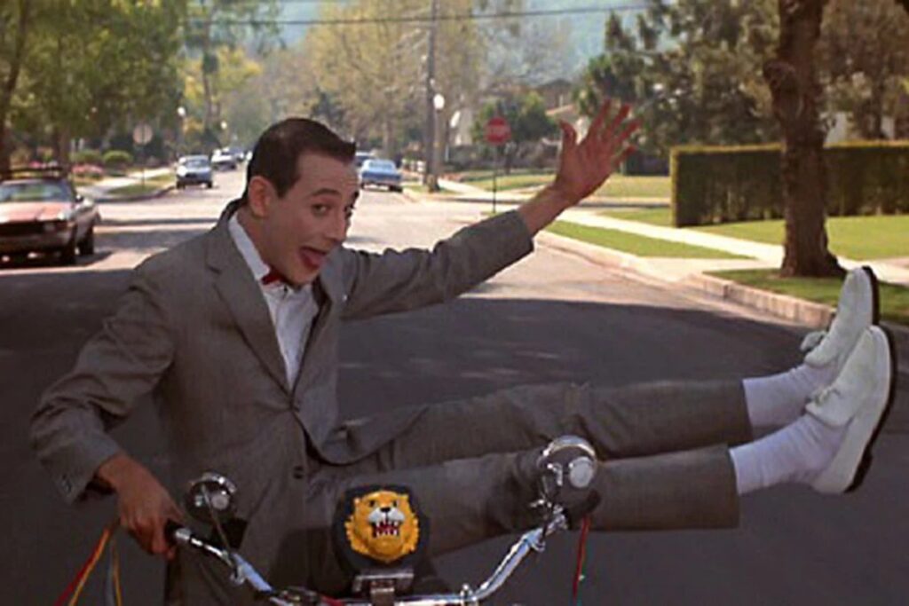 goofy man in tight fitting gray suit balancing sideways on a bicycle with a silly grin on his face