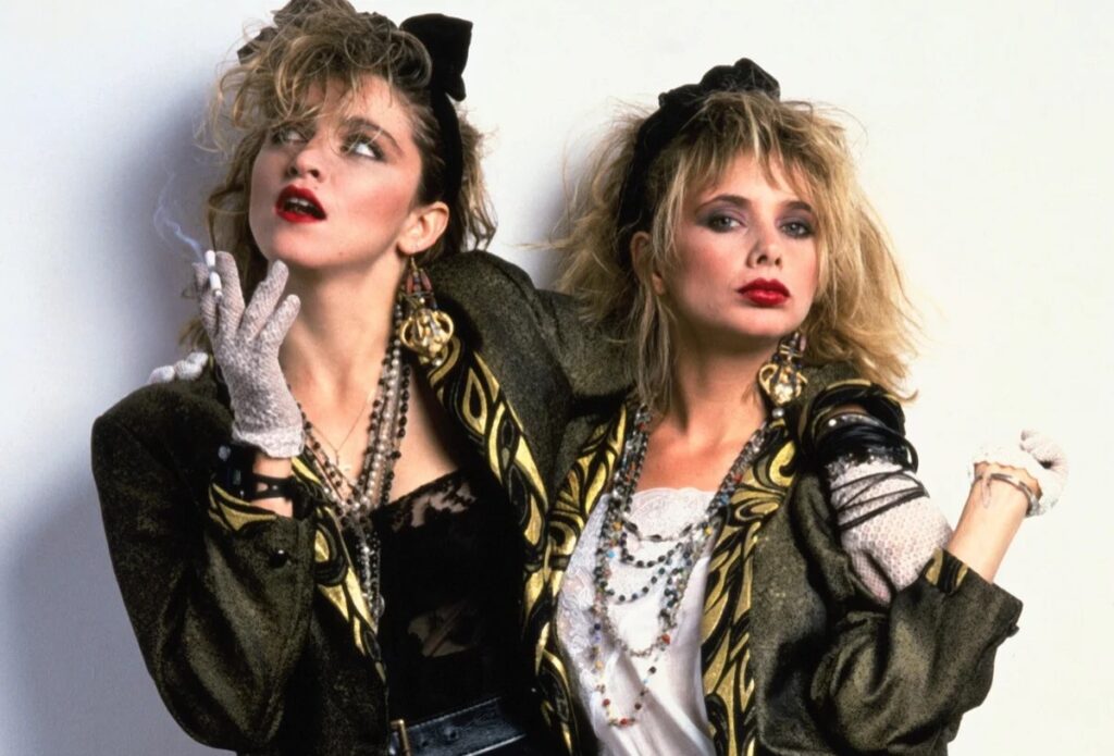 two young women, similar in appearance, dressed in 1980s New Wave outfits and striking a sultry pose for the camera