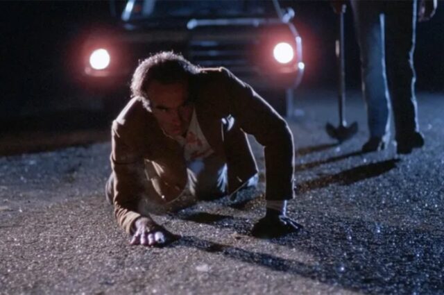 man in tan suit crawling on the pavement at night in front of truck with headlights glaring. Feet of a man holding an axe is off to the right.