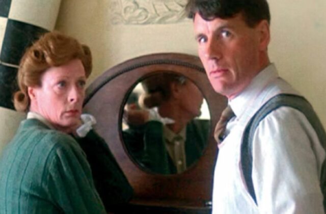 a woman in a green dress and tight bun looking at a nervous man in white shirt and suspenders as he looks over his shoulder.