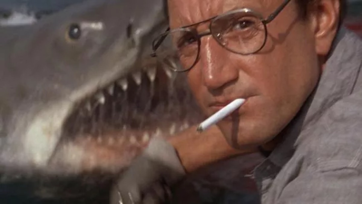 man in glasses with cigarette in his mouth looking away as a shark looms up behind him.