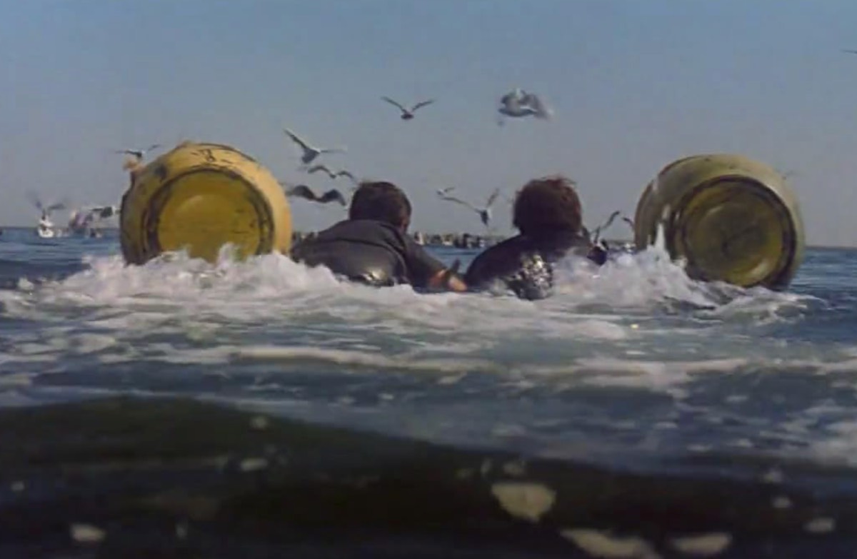 shot of the backs of two men in the water, kicking and holding on to a makeshift buoyancy apparatus