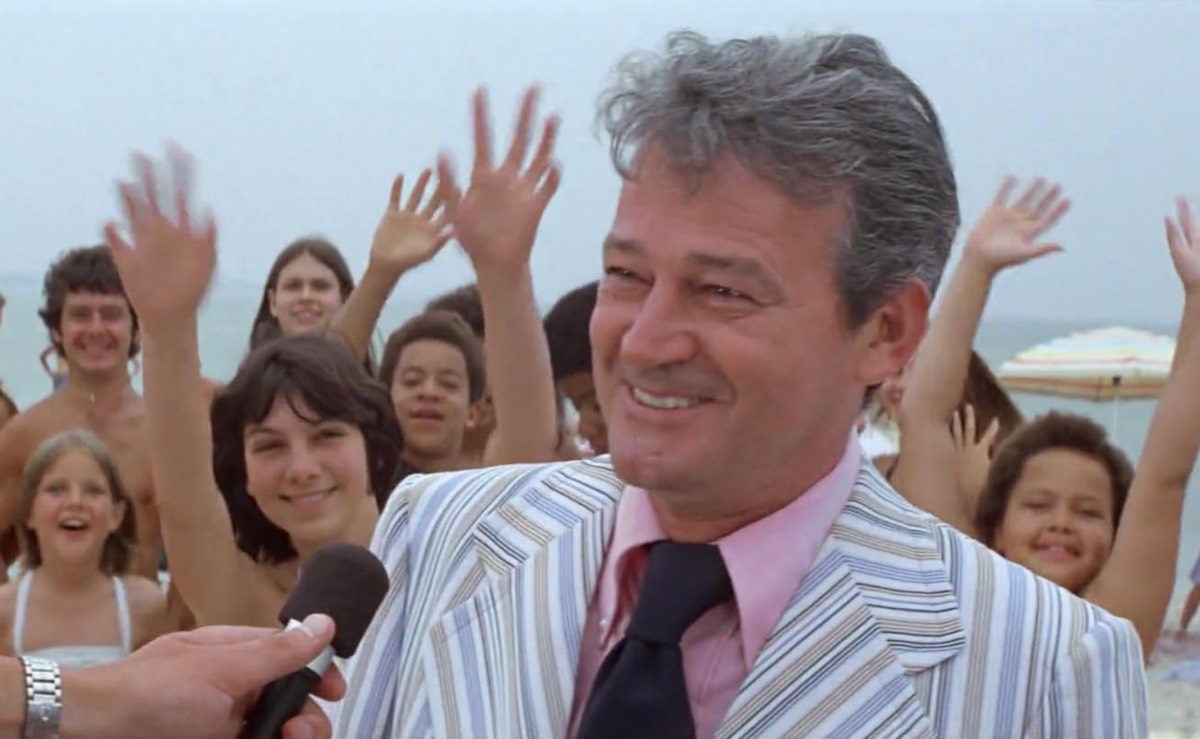 Smug gray haired man in a striped suut and purple tie smiling into camera as residents wave hands behind him