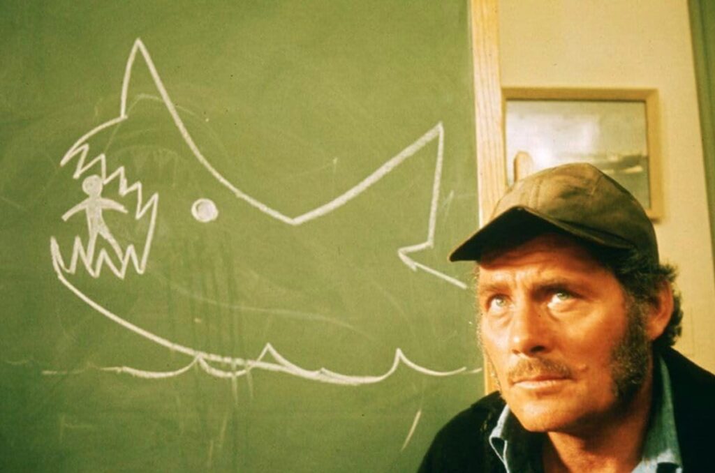 man in fishing cap sitting by blackboard with a crude drawing of a shark on it.