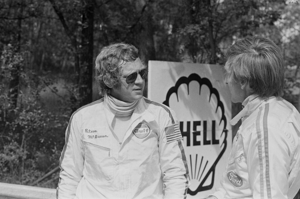 A black and white photo of Steve McQueen during the filming of Le Mans, talking to someone who may be Derek Bell.