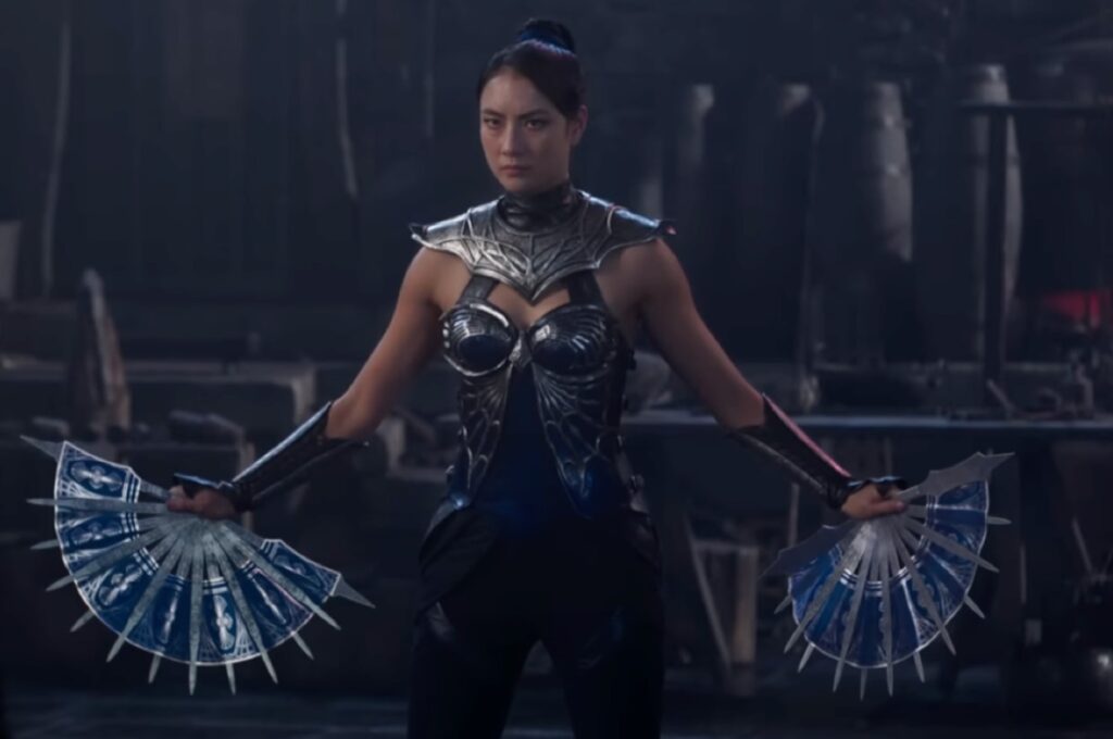 Dark haired woman in battle armor holding two very sharp fans
