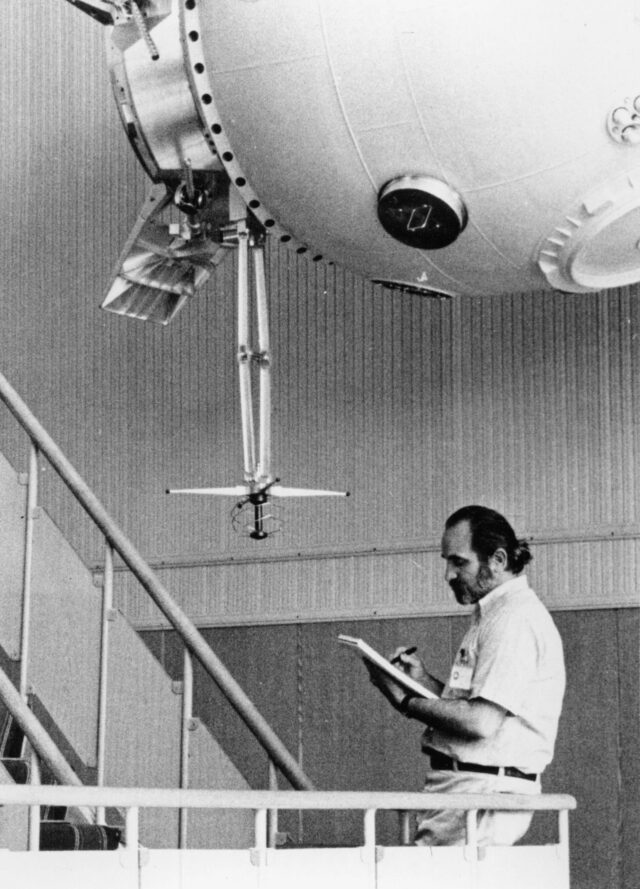 black and white photo of a man in a white shirt holding a pad of paper and standing underneath a mock spacecraft
