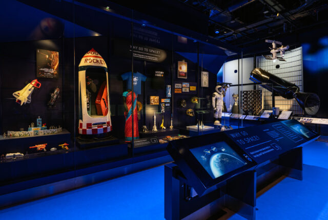 a black and white astronaut's pressure suit and other space artifacts are displayed behind glass in a museum gallery with blue flooring and walls