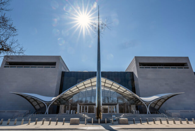 the exterior entrance to a building with a tall, spike-like silver sculpture standing front and center