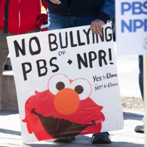 People at a rally urge Congress to protect funding for public broadcasters. A man carries a sign that has a picture of Sesame Street character Elmo and says, "No bullying of PBS and NPR: Yes to Elmo, No to Elon." Another sign says, "Defend PBS and NPR."