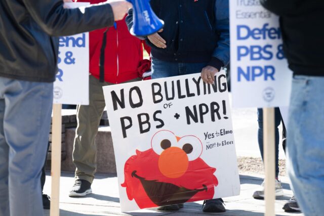 People at a rally urge Congress to protect funding for public broadcasters. A man carries a sign that has a picture of Sesame Street character Elmo and says, "No bullying of PBS and NPR: Yes to Elmo, No to Elon." Another sign says, "Defend PBS and NPR."