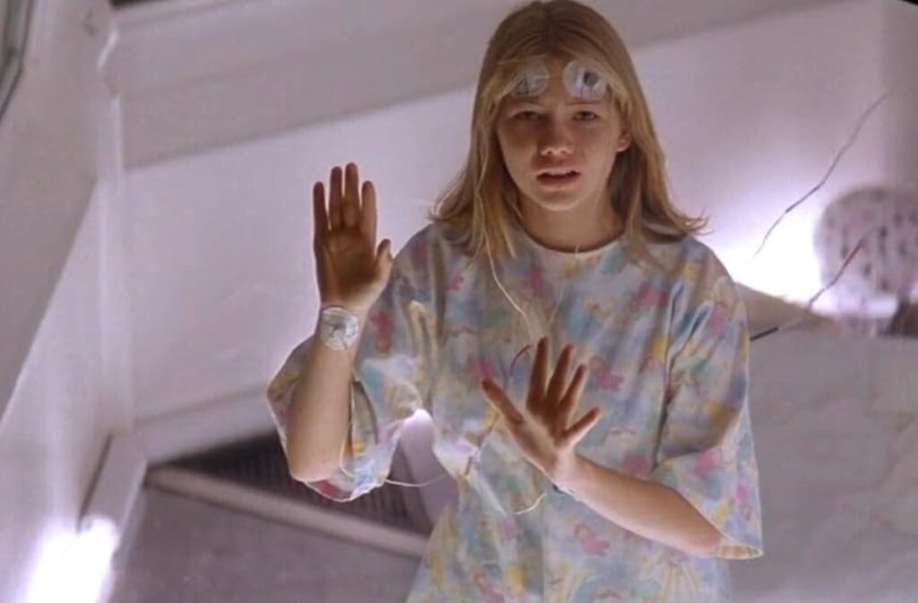 young blonde girl in hospital gown looking through a glass barrier with both hands on the glass.